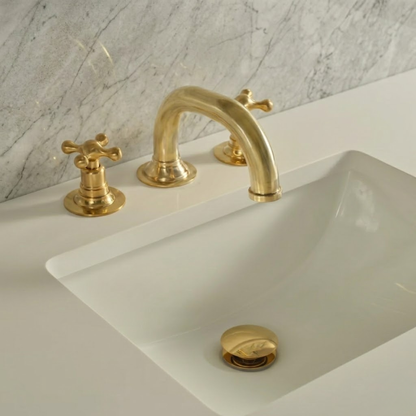Solid unlacquered brass widespread bathroom faucet with cross handles and matching brass pop-up drain installed on a white sink with marble wall background