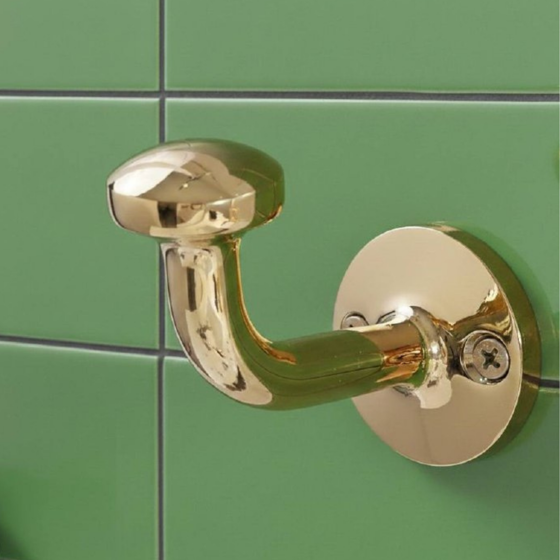 Close-up of an unlacquered solid brass wall hook mounted on green ceramic tile, featuring a vintage curved design and living brass finish.