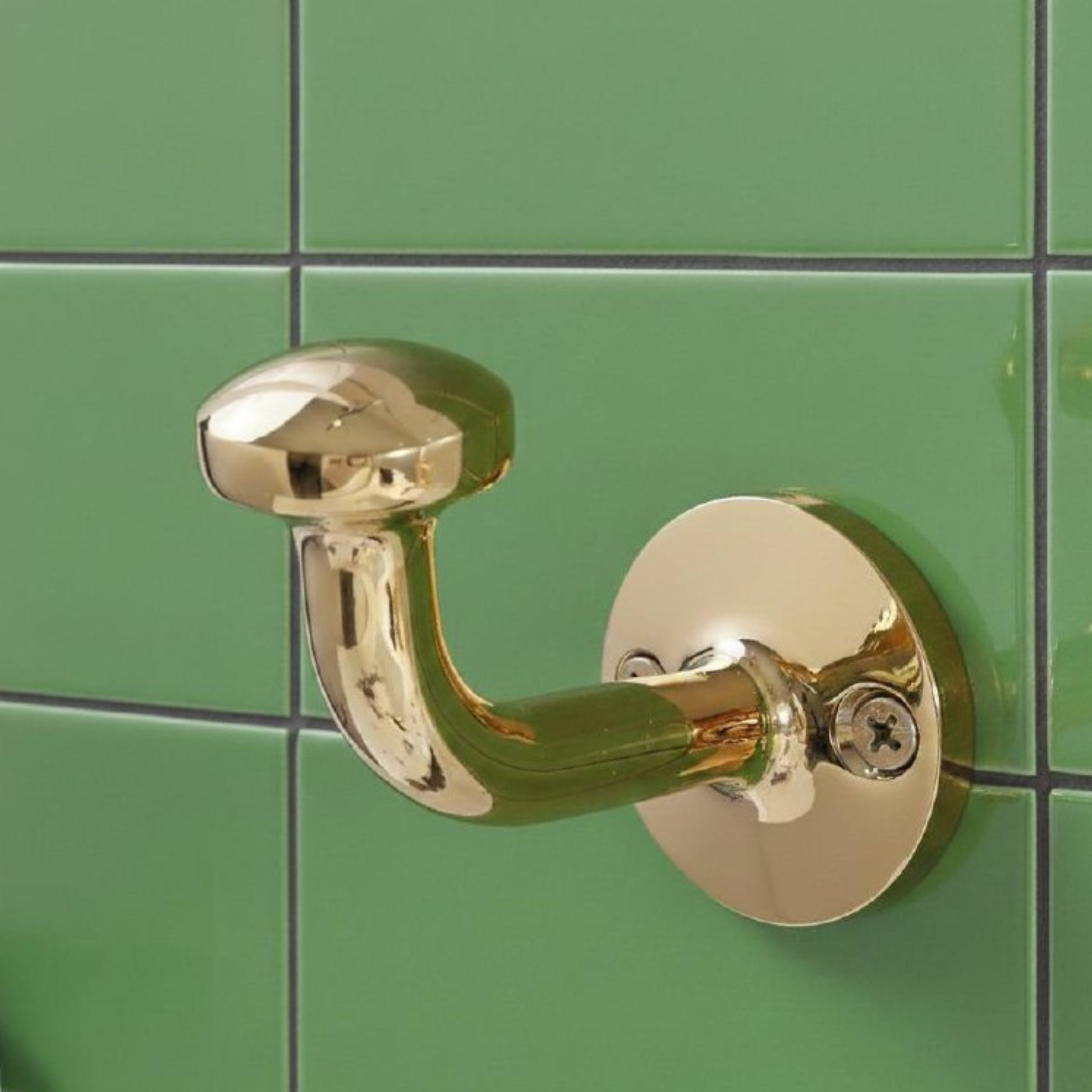 Front view of a vintage-style solid brass wall hook with unlacquered finish, mounted on green tiled wall for bathroom or entryway use.