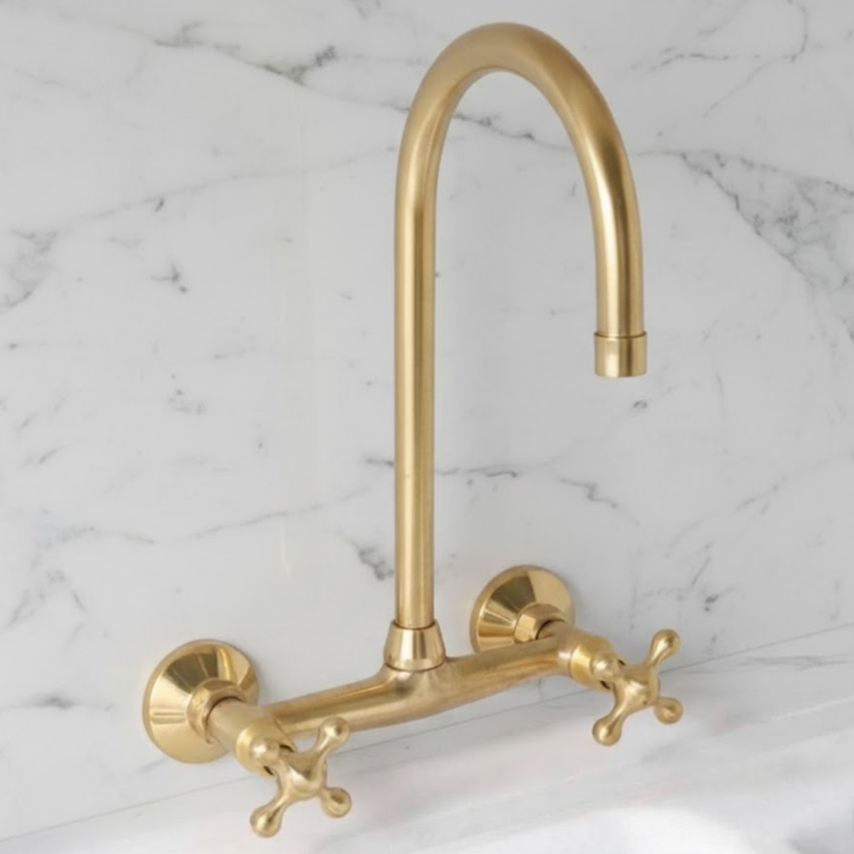 Unlacquered solid brass wall-mounted bridge kitchen faucet with high-arc spout and cross handles on white marble backsplash