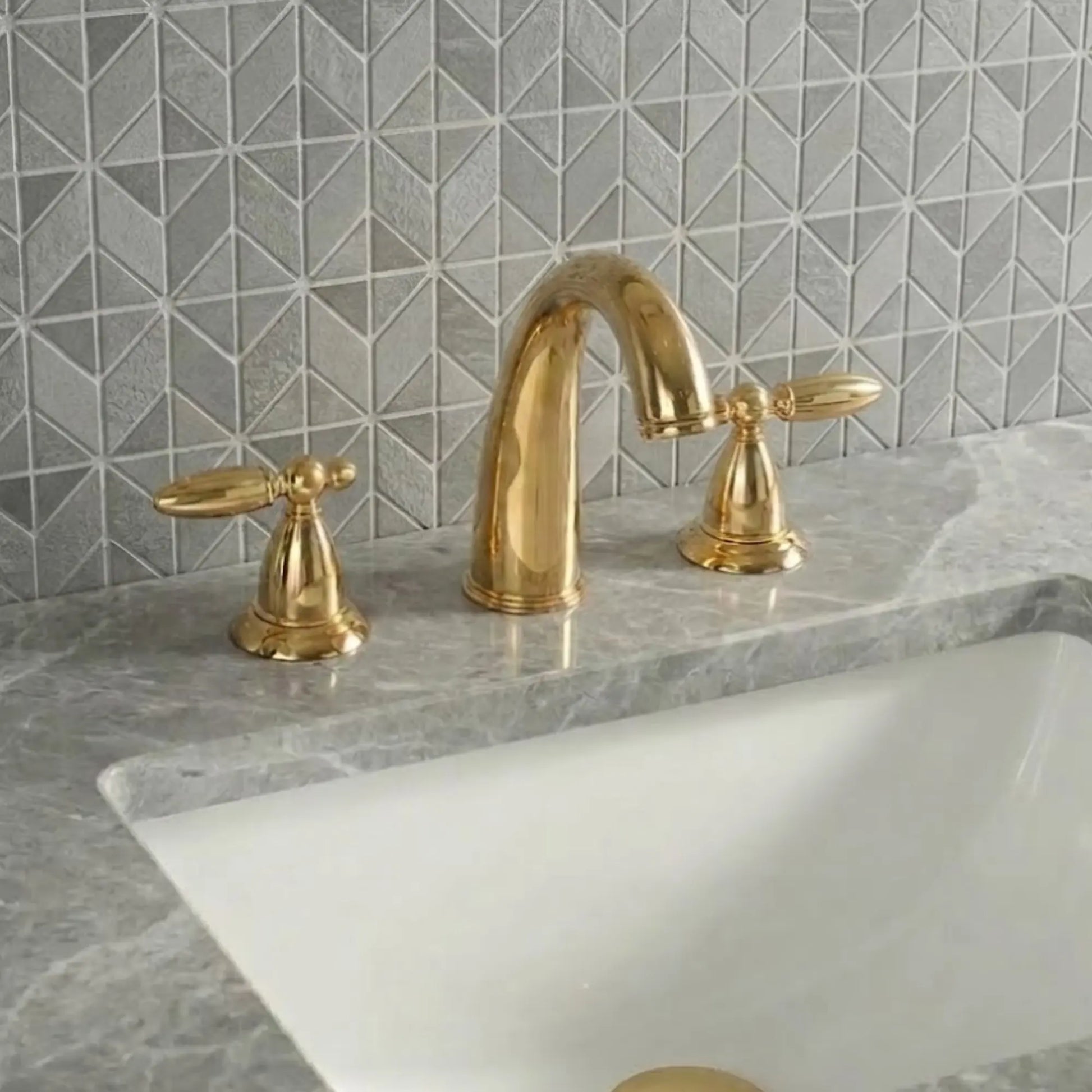Sella unlacquered brass bathroom faucet side angle view highlighting solid brass cross handle detail with gooseneck spout and geometric tile backsplash