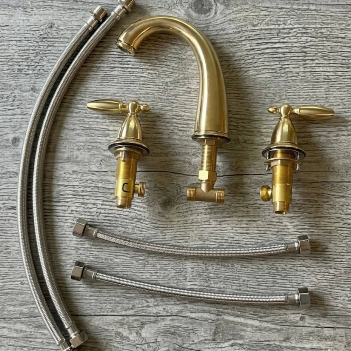 Sella unlacquered brass widespread faucet complete set showing gooseneck spout, two cross handles with valve bodies, and braided stainless steel supply hoses laid flat on wood surface