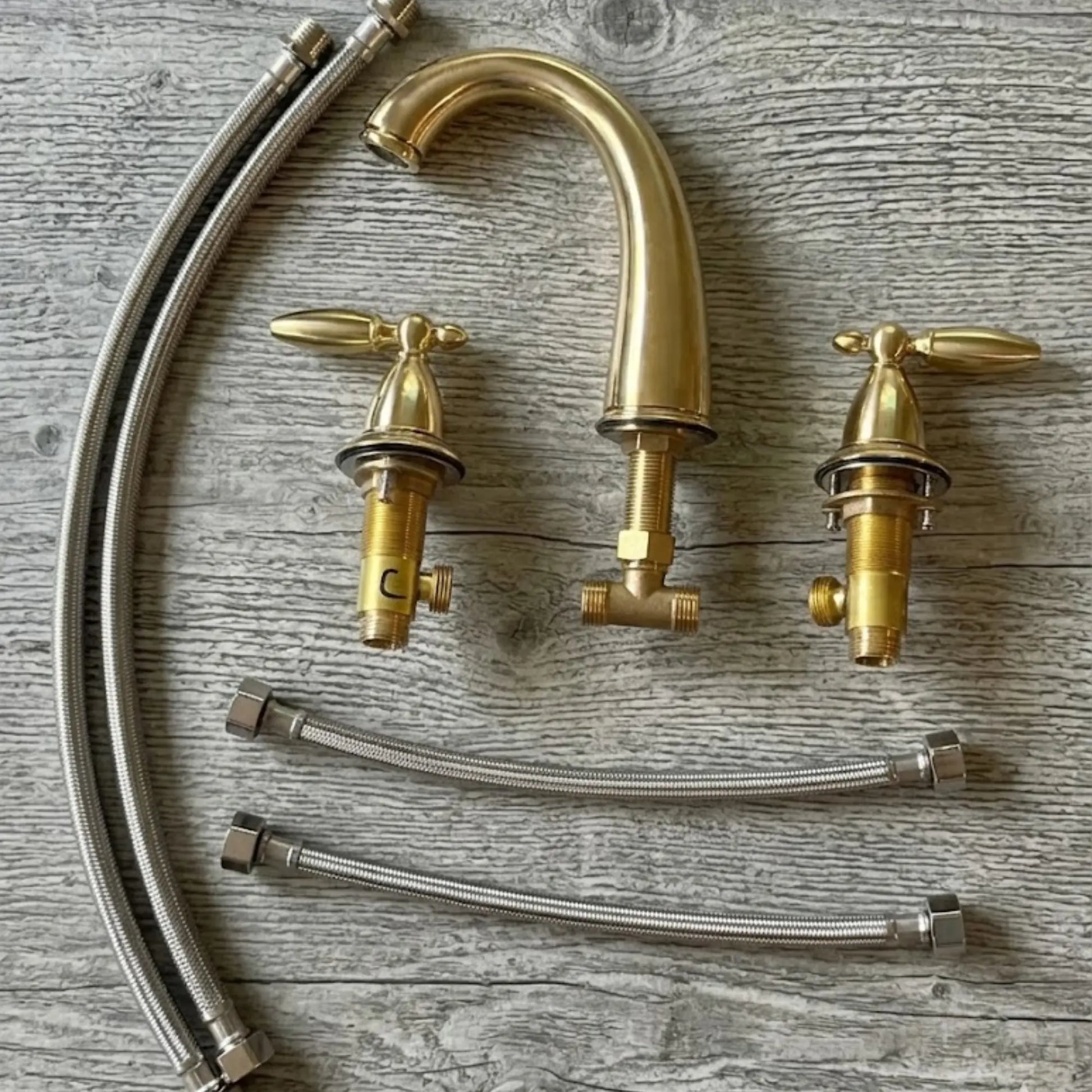 Sella unlacquered brass widespread faucet complete set showing gooseneck spout, two cross handles with valve bodies, and braided stainless steel supply hoses laid flat on wood surface