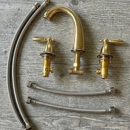 Sella unlacquered brass widespread faucet complete set showing gooseneck spout, two cross handles with valve bodies, and braided stainless steel supply hoses laid flat on wood surface