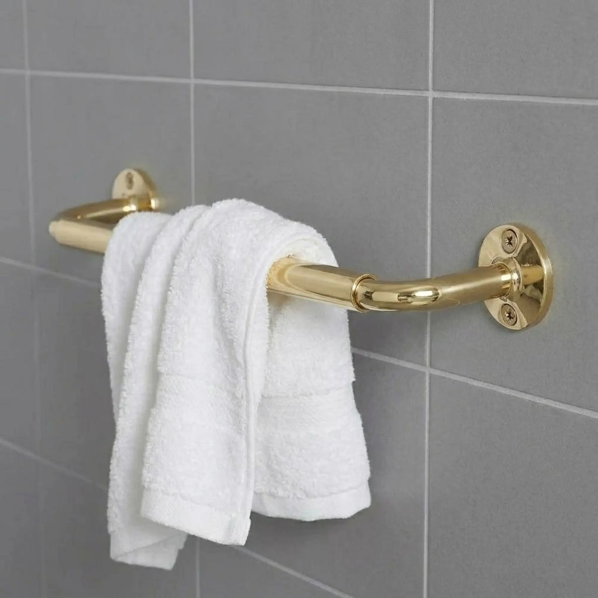 Detailed view of a luxury brass bathroom rail with a white towel, highlighting the sturdy wall-mount flanges and polished gold finish against a grey tiled backdrop.