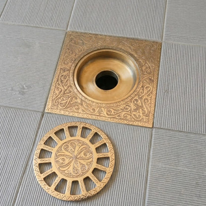 Engraved brass square shower floor drain with circular grate removed, showing floor mount installation on white tile