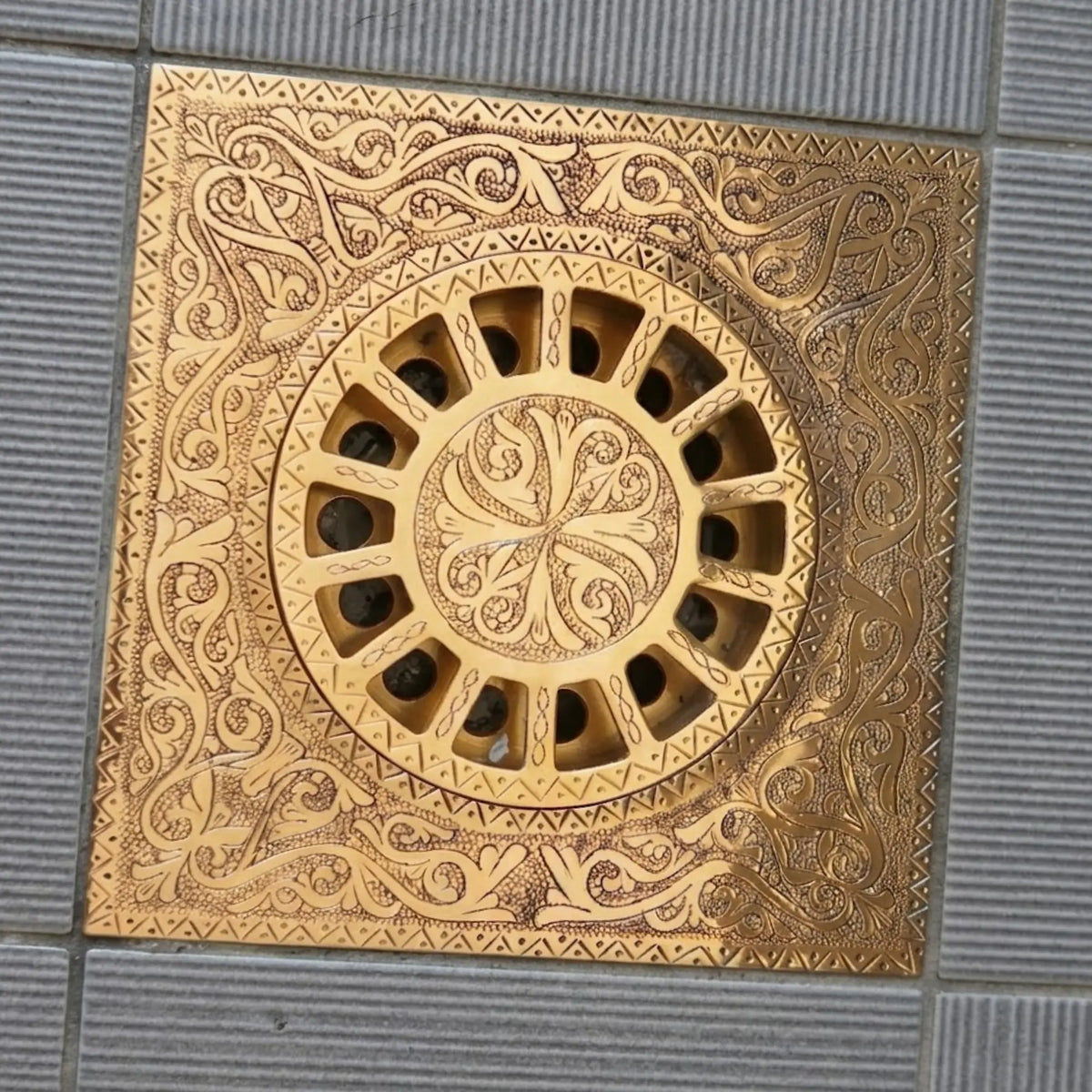 Engraved unlacquered brass square shower floor drain with hand-carved scrollwork pattern installed on tiled floor
