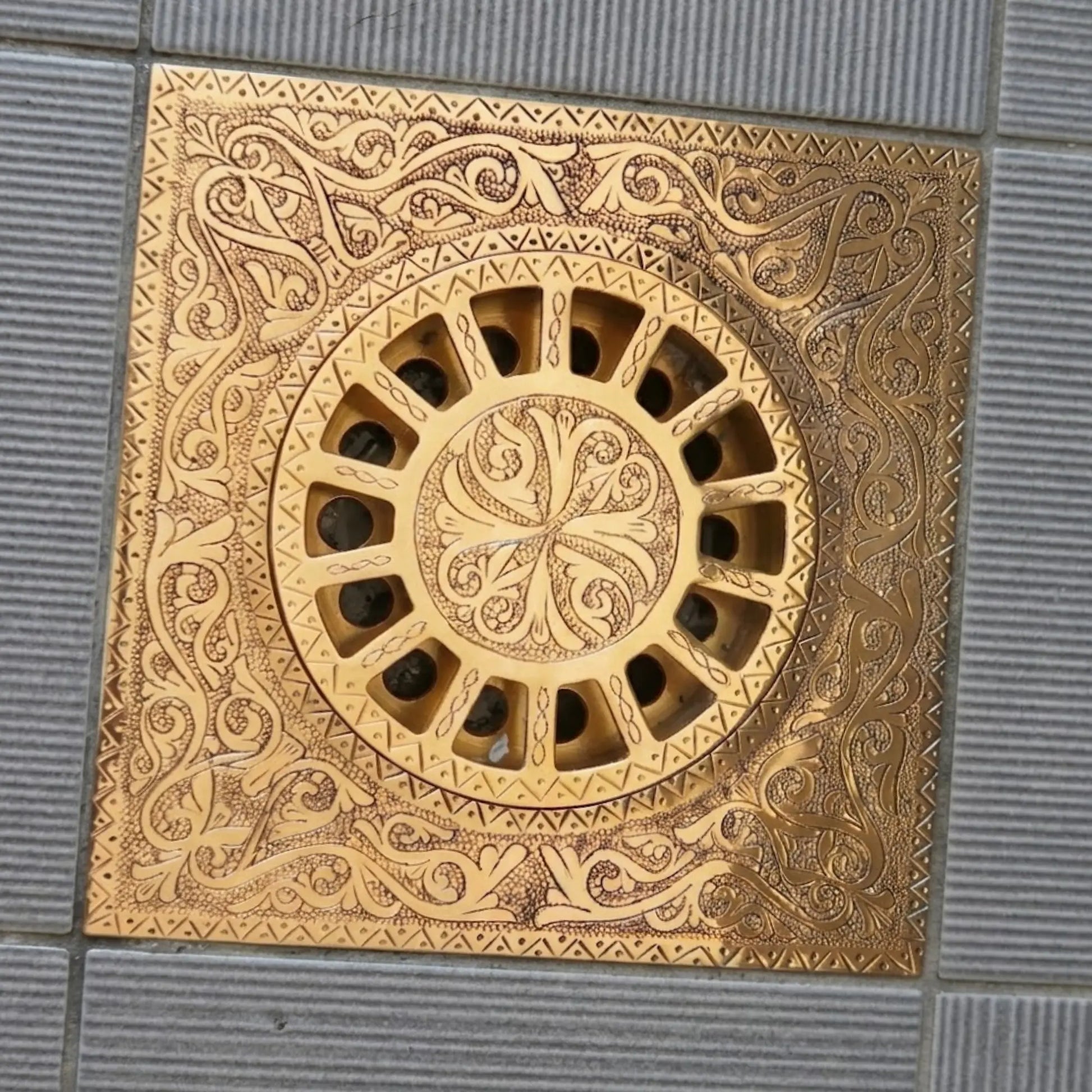 Engraved unlacquered brass square shower floor drain with hand-carved scrollwork pattern installed on tiled floor