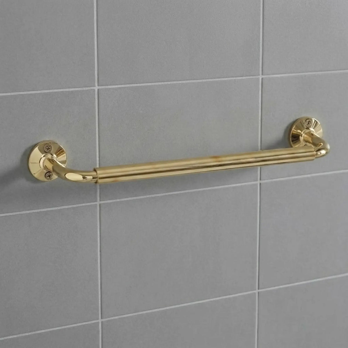 Unlacquered brass wall-mounted grab bar installed on grey subway tiles, featuring traditional round decorative flanges and exposed screws.