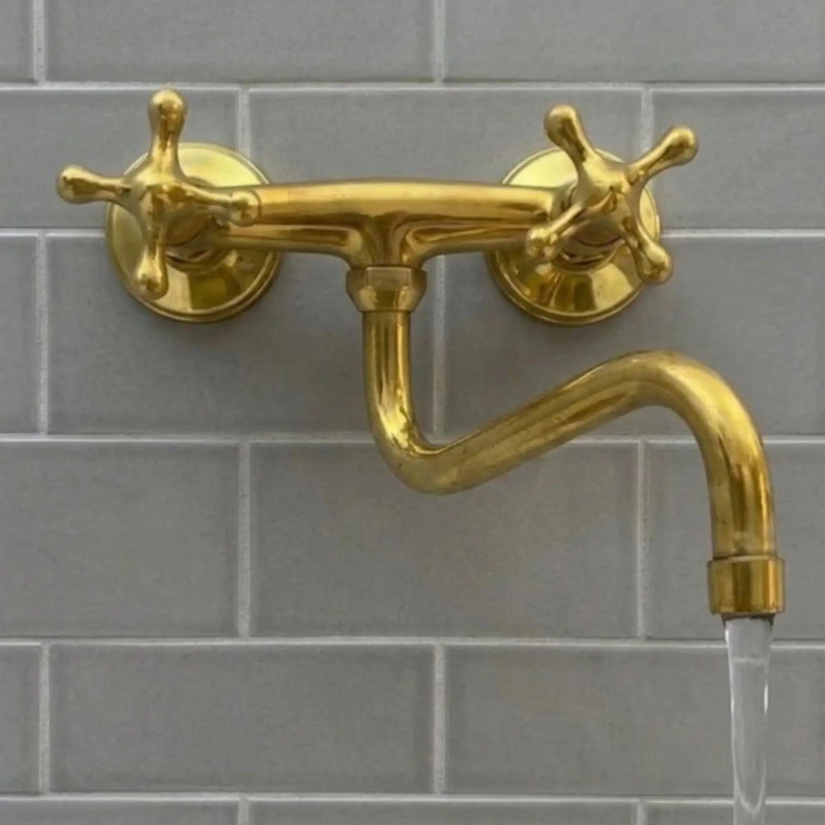 A close-up of a brass wall-mounted bridge faucet with cross handles and an S-spout, featuring a steady, crystal-clear laminar water flow against a grey subway tile backsplash.