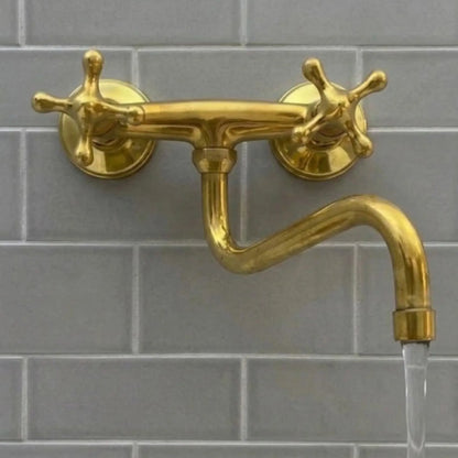 A close-up of a brass wall-mounted bridge faucet with cross handles and an S-spout, featuring a steady, crystal-clear laminar water flow against a grey subway tile backsplash.