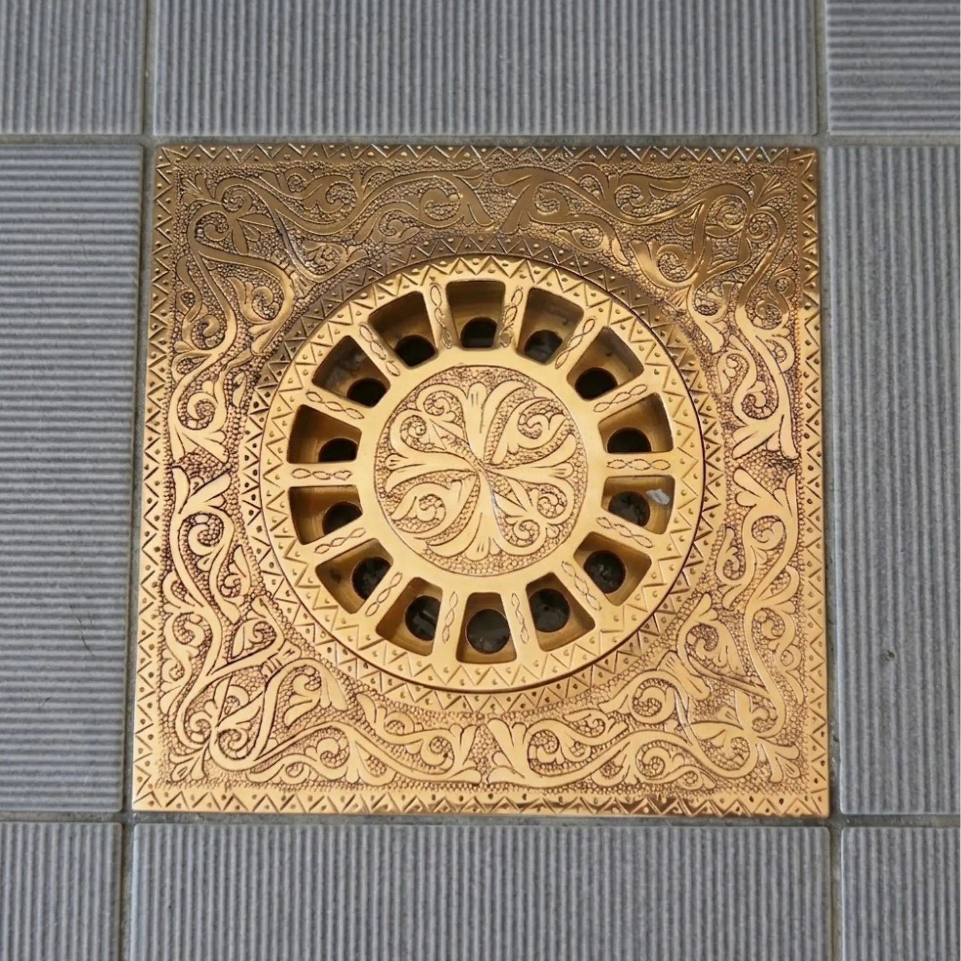 Solid brass square floor drain with decorative engraved pattern and removable circular grate on ceramic tile