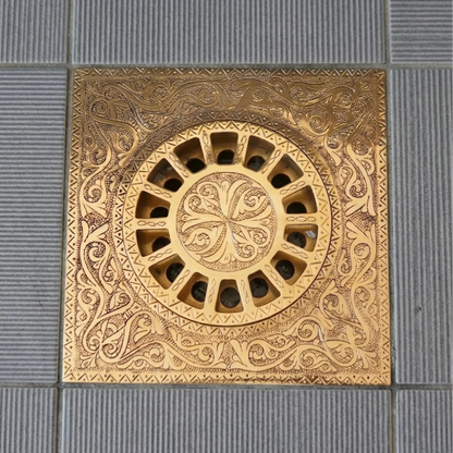 Solid brass square floor drain with decorative engraved pattern and removable circular grate on ceramic tile