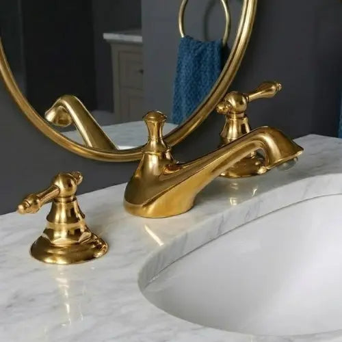 An unlacquered brass widespread bathroom faucet with traditional levers on a white marble countertop next to an under-mount sink, with a round mirror reflection.