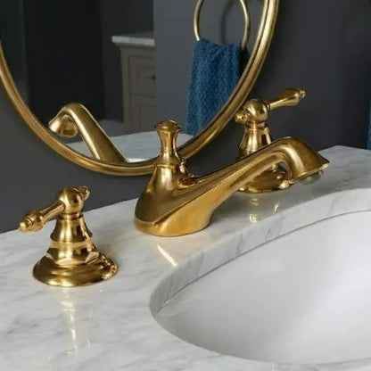 An unlacquered brass widespread bathroom faucet with traditional levers on a white marble countertop next to an under-mount sink, with a round mirror reflection.