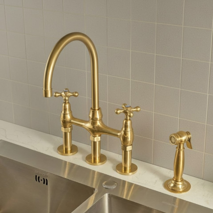 Unlacquered brass 3-hole bridge kitchen faucet with cross handles and side sprayer installed on a white countertop with a stainless steel sink.