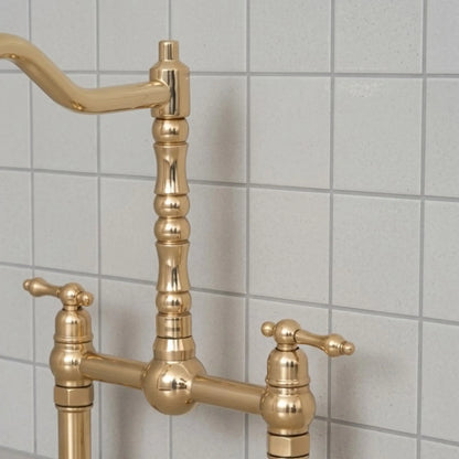 Unlacquered brass bridge kitchen faucet with cross handles and high arc spout installed against tiled backsplash