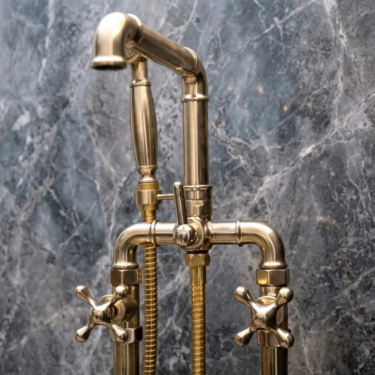 Close-up of unlacquered brass freestanding bathtub faucet with cross handles and handheld shower on gray marble wall