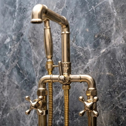 Close-up of unlacquered brass freestanding bathtub faucet with cross handles and handheld shower on gray marble wall