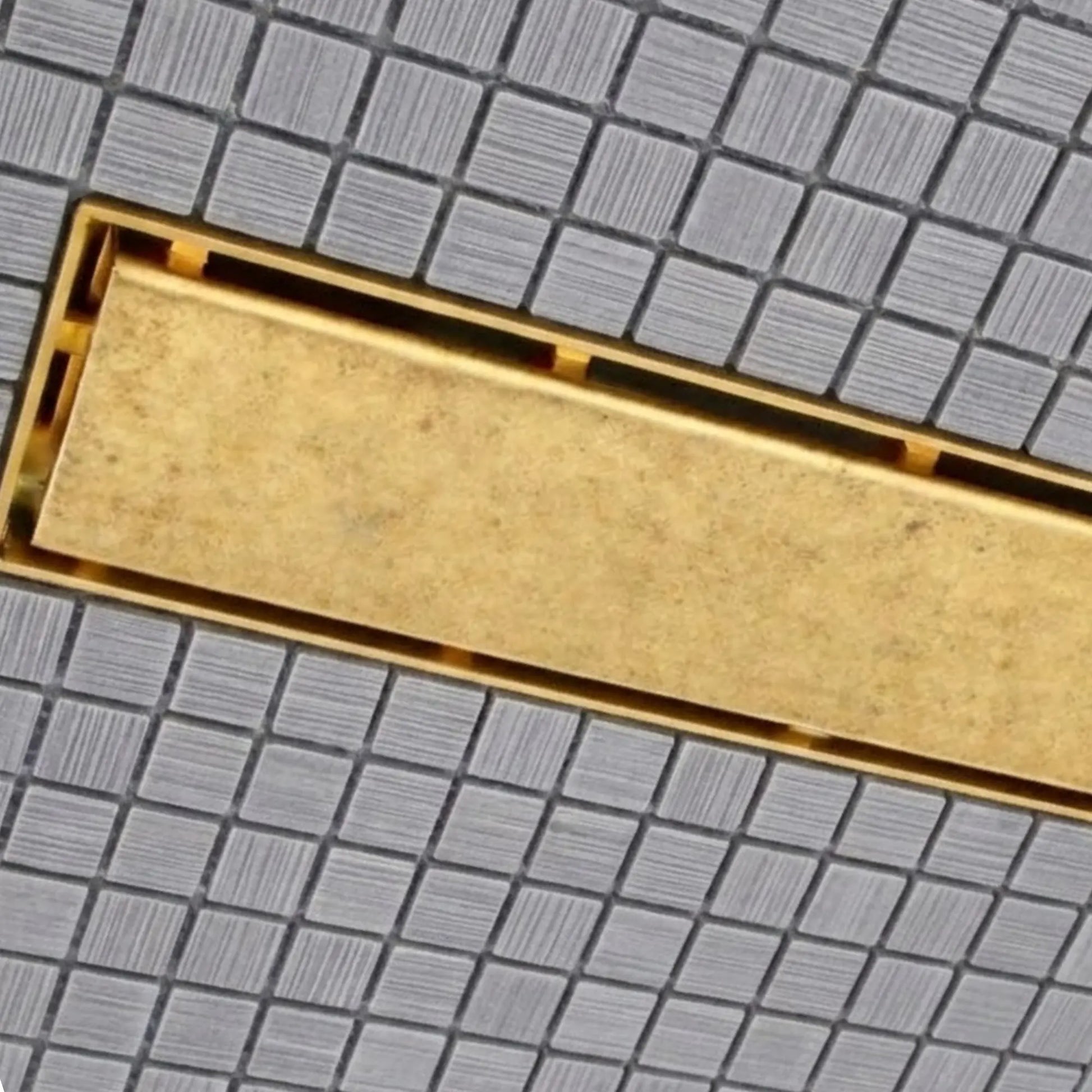 Close-up of unlacquered brass linear shower drain grate showing natural patina texture on mosaic tile – BrassOna