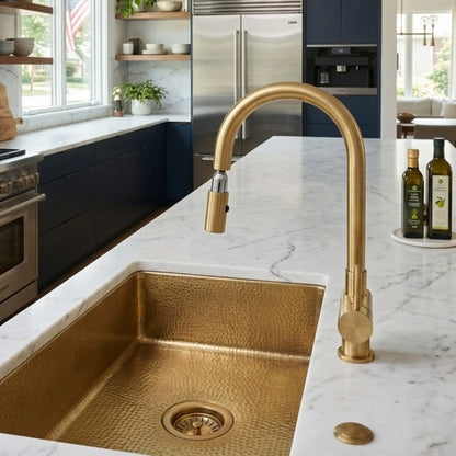 Unlacquered brass pull down kitchen faucet with high arc spout over hammered brass farmhouse sink on white marble countertop