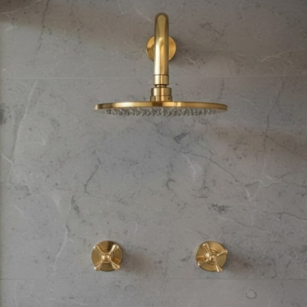 Front view of a round unlacquered brass rainfall shower head and two cross-handle control valves symmetrically mounted on a gray stone-textured wall.