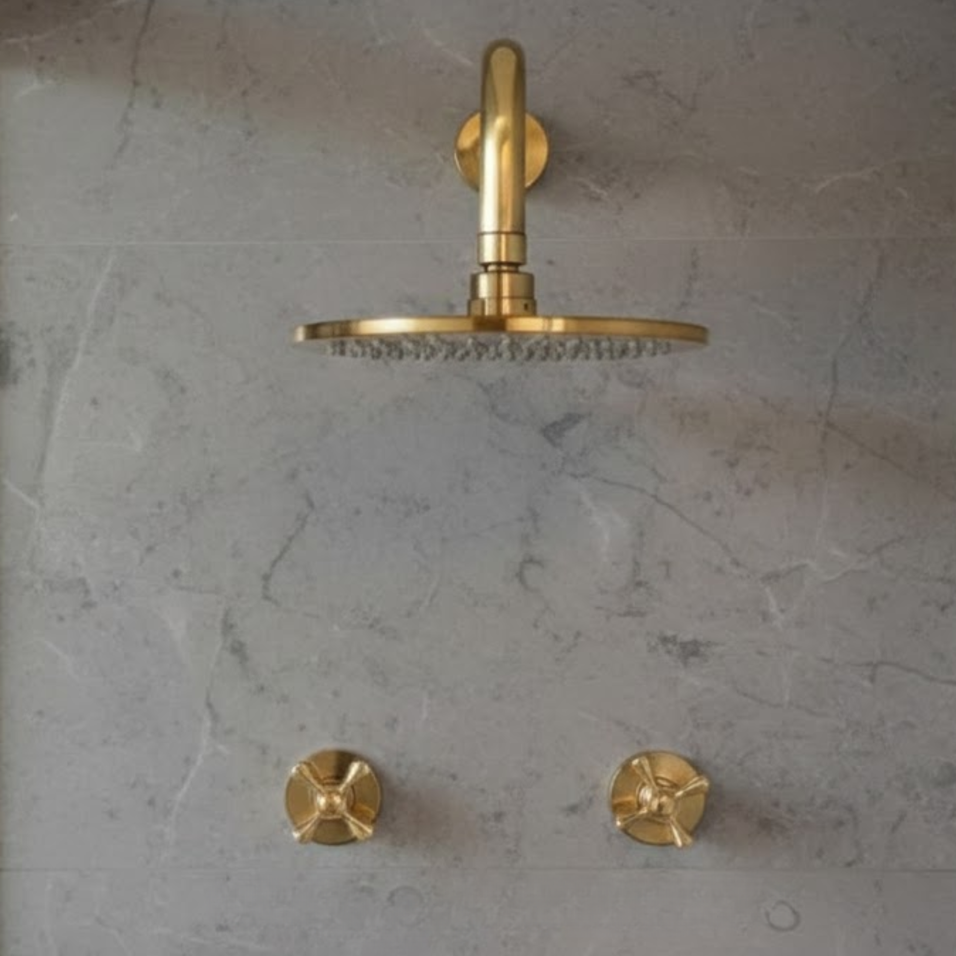 Front view of a round unlacquered brass rainfall shower head and two cross-handle control valves symmetrically mounted on a gray stone-textured wall.
