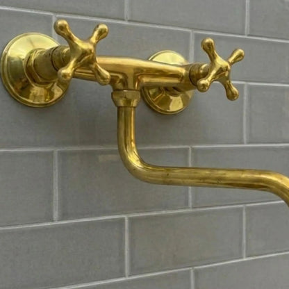 Industrial-luxury unlacquered brass wall-mounted bridge faucet with articulating S-spout and cross handles on a grey subway tile backsplash.