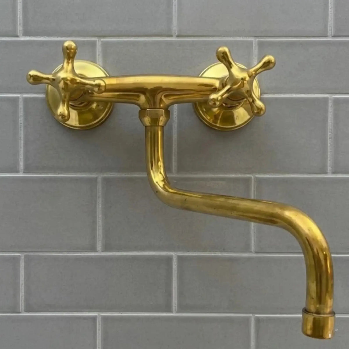 A luxury industrial unlacquered brass wall-mounted bridge faucet with cross handles and an articulating S-spout, set against a minimalist grey subway tile backsplash.