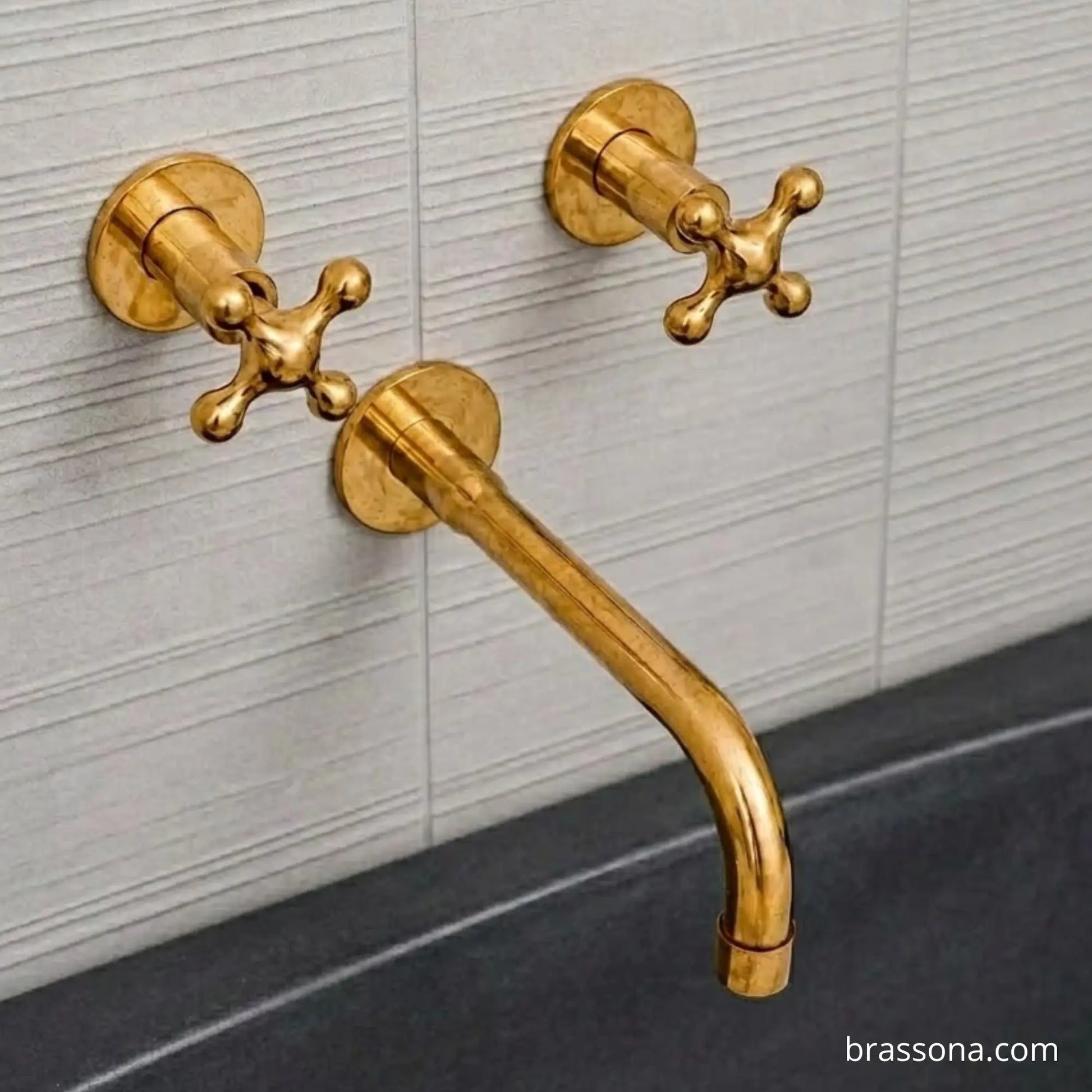 Gold brass wall-mounted bathroom faucet set with two cross handles and a curved spout, installed against a light gray tiled wall above a dark basin.