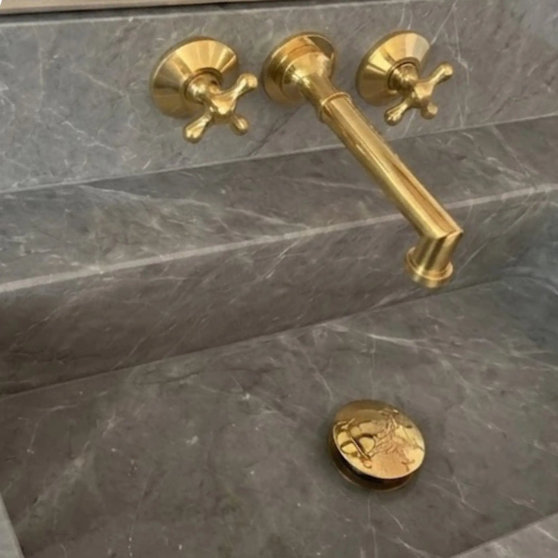 Close up of unlacquered brass wall mount bathroom faucet with cross handles on grey marble sink