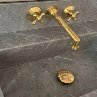 Close up of unlacquered brass wall mount bathroom faucet with cross handles on grey marble sink