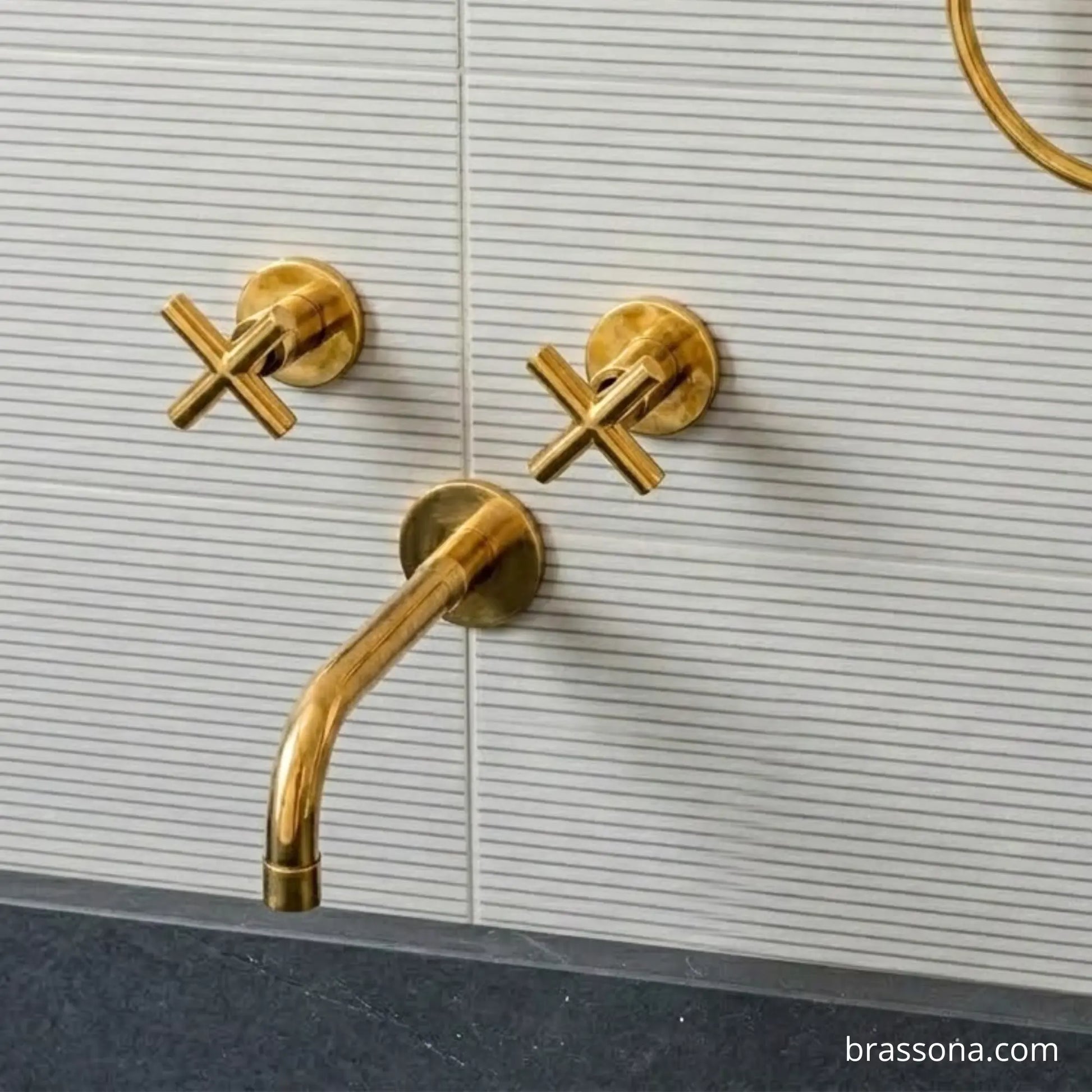 Gold brass wall-mounted bathroom faucet and two cross-handle valves on a white ribbed tile wall above a dark countertop.