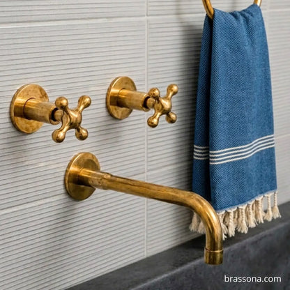 Gold brass wall-mounted bathroom fixtures including two cross-handle valves and a curved spout, shown against a light gray ribbed tile wall with a blue towel hanging on a gold ring.