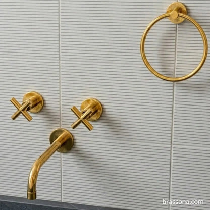 Gold brass bathroom fixtures including a curved faucet spout, two cross-handle valves, and a round towel ring mounted on a white ribbed tile wall. A small Brassona watermark is visible in the bottom right corner.