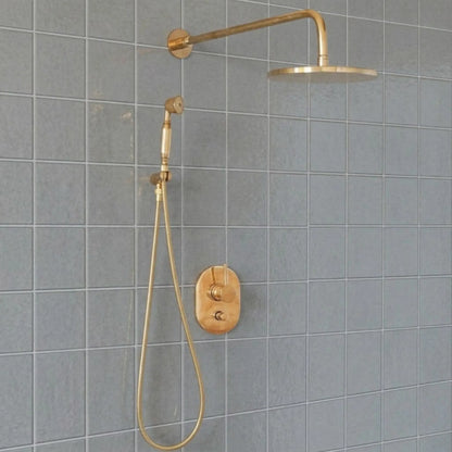 Unlacquered brass wall mount rain shower system with handheld sprayer and exposed pipe design installed on gray tile bathroom wall