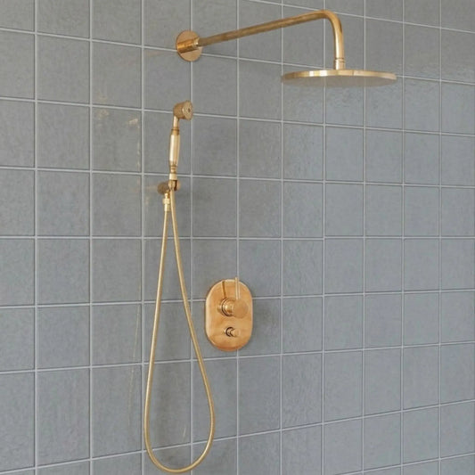 Unlacquered brass wall mount rain shower system with handheld sprayer and exposed pipe design installed on gray tile bathroom wall