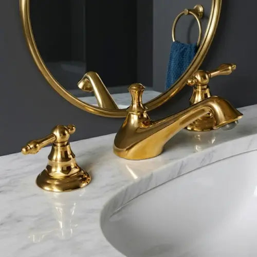 Luxury unlacquered brass widespread bathroom faucet with traditional lever handles, mounted on a white marble vanity countertop against a dark gray wall with a matching round brass mirror.