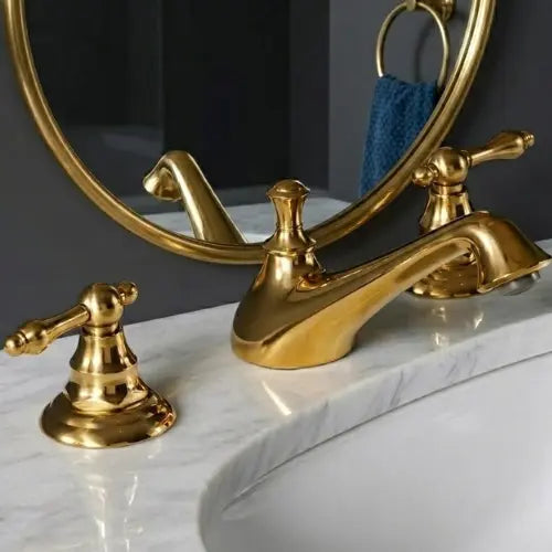 Close-up view of an unlacquered brass widespread bathroom faucet with classical lever handles, installed on a white marble vanity top next to an under-mount sink basin.