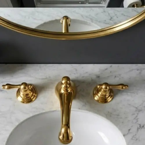Top-down overhead view of an unlacquered brass widespread bathroom faucet with dual lever handles, installed on a white Carrara marble vanity countertop with an under-mount sink basin.