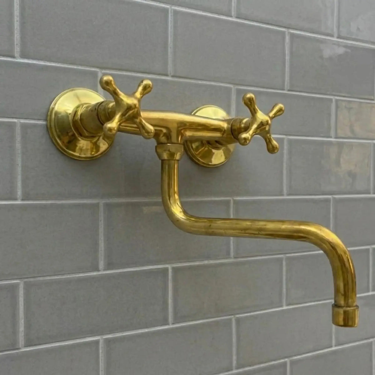 an unlacquered brass wall-mounted bridge faucet with cross handles and an articulating S-spout, set against a minimalist grey subway tile backsplash.