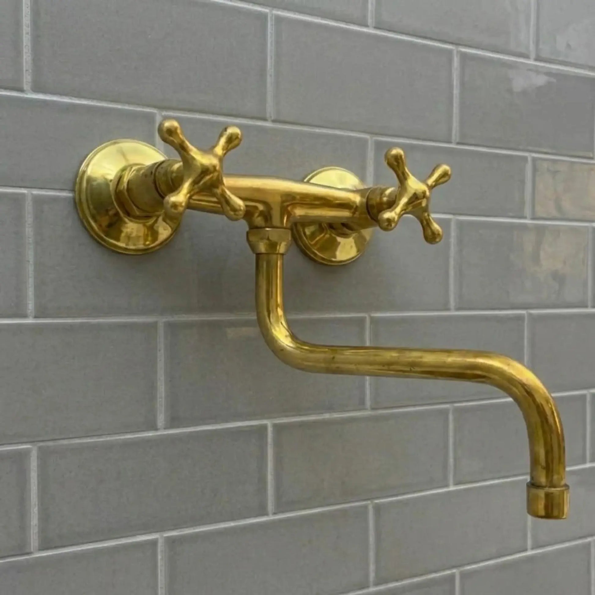 an unlacquered brass wall-mounted bridge faucet with cross handles and an articulating S-spout, set against a minimalist grey subway tile backsplash.