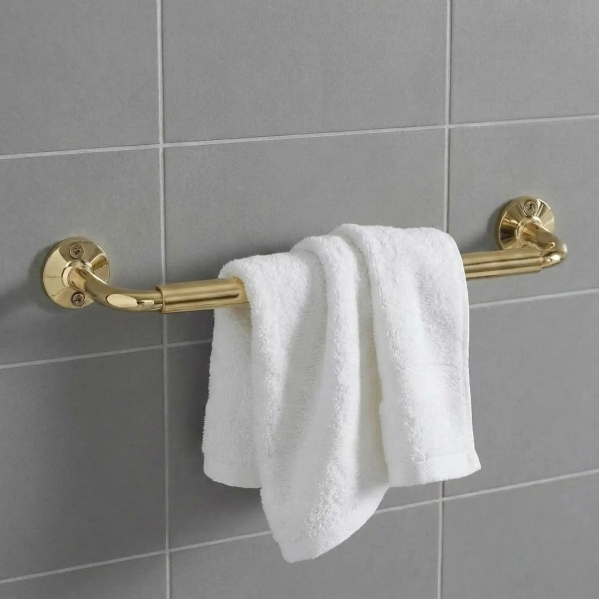 Solid unlacquered brass towel rail mounted on grey tile, featuring a draped white plush cotton towel in a minimalist bathroom setting.